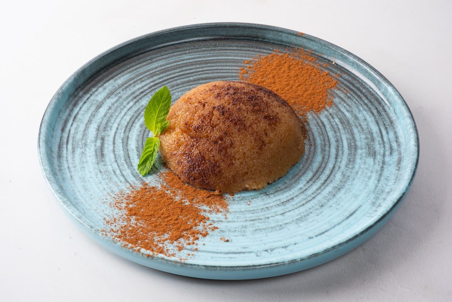 Şef'in İrmik Helvası shaped as a dome on a blue ceramic plate with cinnamon and a mint leaf
